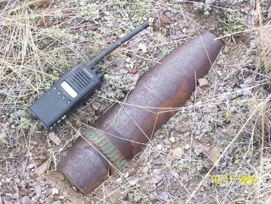 Rusted, half-buried artillery projectile in scrub with a handheld radio for scale